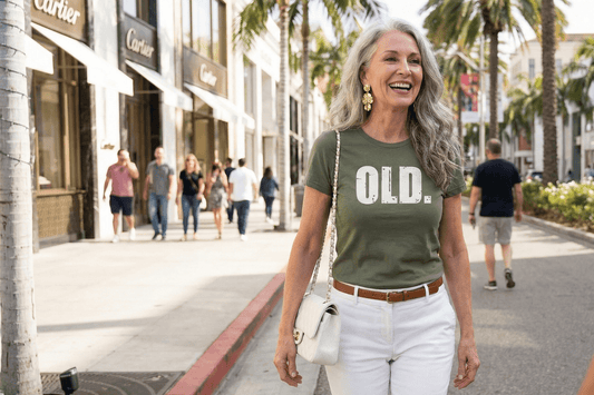 Woman wearing olive green 'OLD.' graphic tee, white pants, walking on palm-lined street