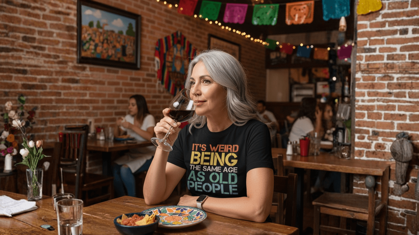 Woman sipping wine in a cozy restaurant, wearing a funny 'It's weird being the same age as old people' tee.